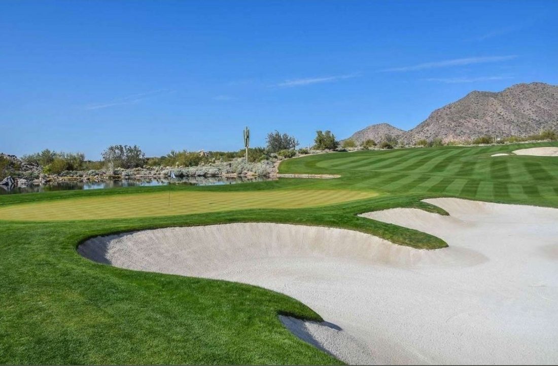 A Silverleaf Club green guarded by a large sculpted bunker, water behind the green, a lone saguaro and the McDowell Mountains beyond